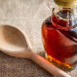 Glass jar of maple syrup next to a wooden spoon.